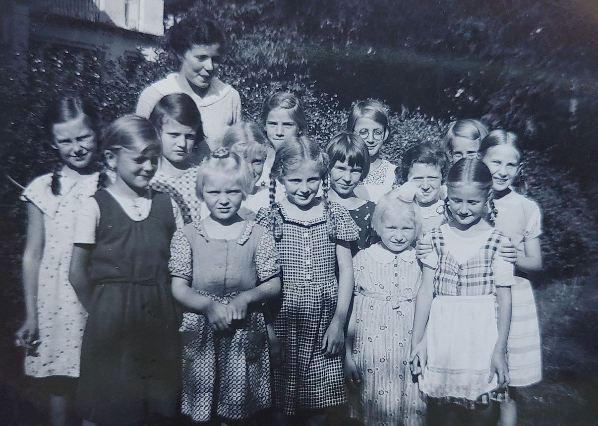 Eine Gruppe von 14 sehr jungen Mädchen, vor einer Hecke im Sonnenlicht stehend. Sie blinzeln gegen die Sonne an, nicht alle lächeln in die Kamera, manche schauen grimmig, andere skeptisch. Hinter ihnen steht eine freundlich wirkende Erzieherin.