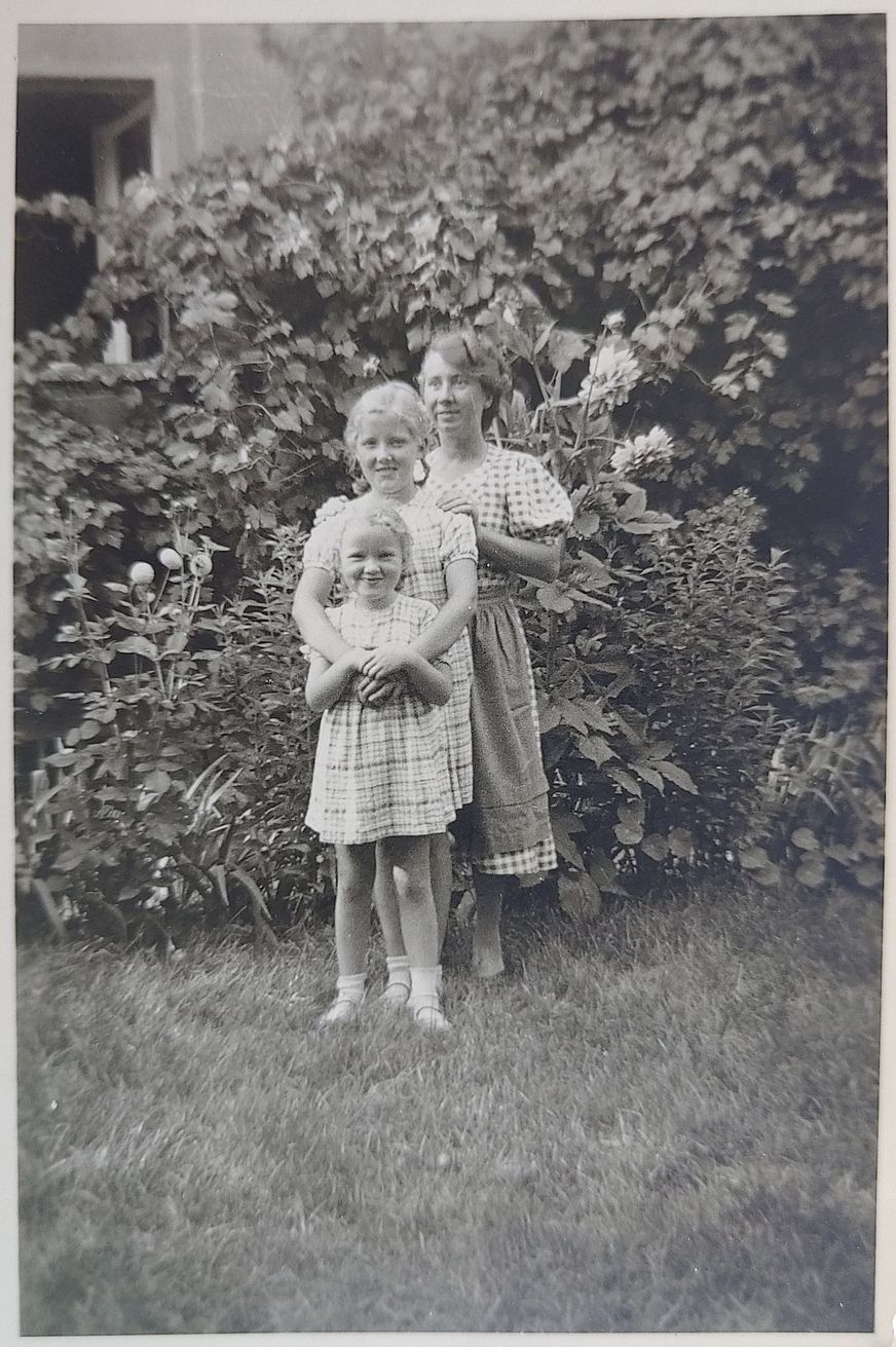 Drei Personen stehen auf einem dicht gewachsenen Rasen, vor einer Hecke, die an einer Hauswand hinaufrankt. Ganz vorn steht ein strahlendes junges Mädchen, hinter ihr ein etwas älteres Mädchen, die Arme über die Schultern der Kleinen vor ihr gelegt. Direkt hinter ihnen steht eine erwachsene Frau, nur einen halben Kopf größer als das Mädchen. Sie hat die Hand auf ihrer Schulter. Alle lächeln und stehen sehr dicht beieinander.