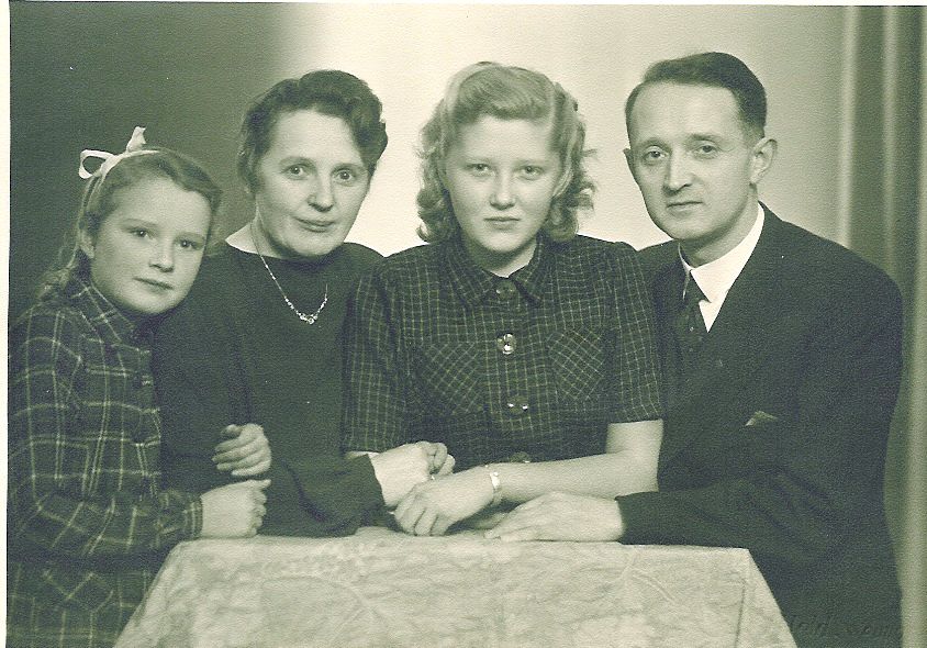 Eine blonde junge Frau sitzt zwischen einem Mann und einer Frau, ein junges Mädchen drückt sich von rechts an die Frau. Sie sitzen alle eng aneinander gedrückt an einem Tisch, die Arme miteinander verschränkt. Die junge Frau trägt eine hoch geschlossene, kurzärmelige dunkle Bluse und schaut selbstbewusst in die Kamera.