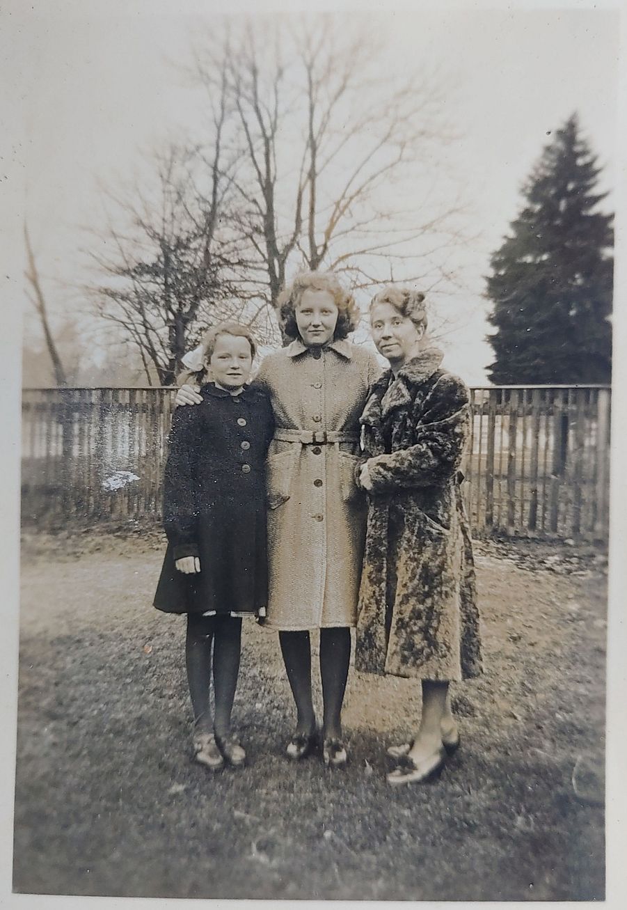 Eine Jugendliche steht zwischen einem jungen Mädchen und einer älteren Frau. Alle tragen Winterkleidung. Sie hat den Arm um die Schultern des Mädchens gelegt. Sie stehen auf einer abgetretenen Wiese, vor einem Zaun, im Hintergrund sind Bäume die kein Laub mehr tragen, der Himmel ist grau und nebelverhangen.