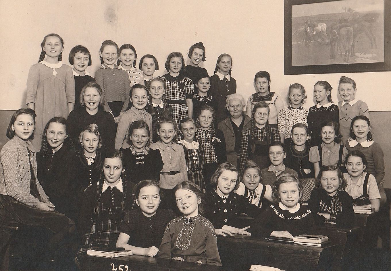 Eine Gruppe von 38 jungen Mädchen, in der Ecke eines Klassenzimmers an Schulbänken sitzend bzw. auf ihnen stehend. Fast alle von ihnen sehen lächelnd und aufgeweckt in die Kamera. In der Mitte der Gruppe sitzt eine weißhaarige, freundlich lächelnde ältere Dame. An der Wand hängt ein großes Gemälde, das eine Szene pausierender Kavalleristen zeigt.