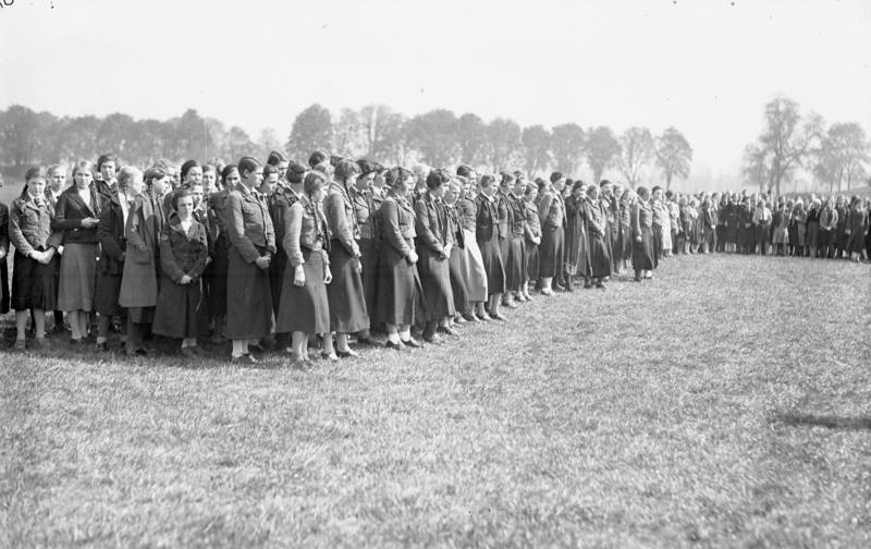 Auf einer Wiese steht eine große Anzahl Mädchen, in Reihen angetreten zum Appell. Sie tragen alle lange, dunkle Röcke und kurze Jacken. Im Hintergrund, am weiter entfernten Rand der Wiese, steht eine einzelne Baumreihe.
