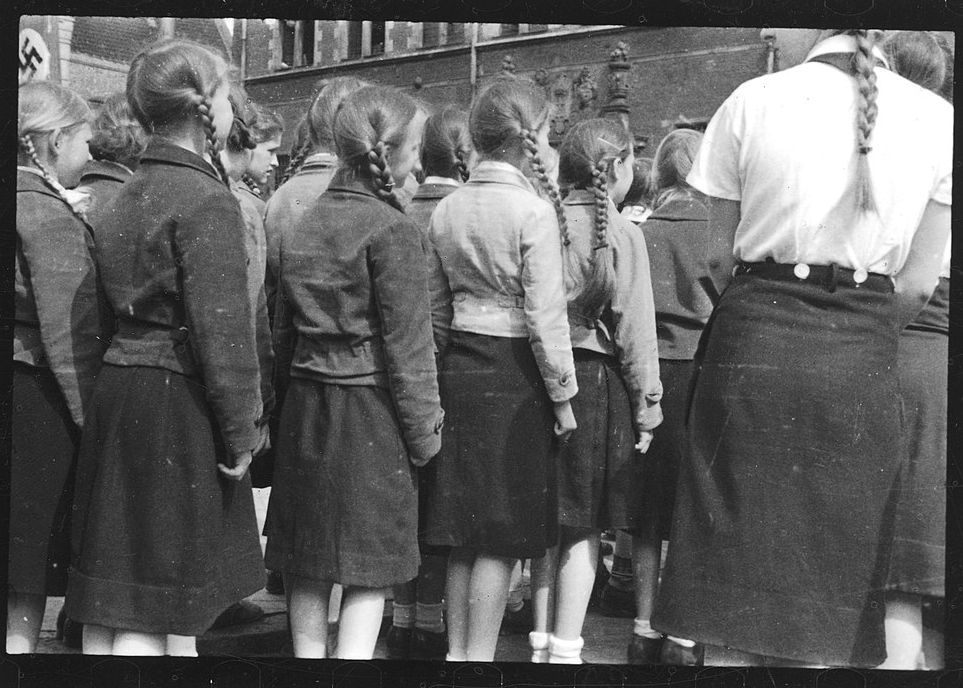 Rund ein Dutzend junger Mädchen, von hinten aufgenommen, in engen Reihen stehend. Alle tragen knielange Röcke und kurze, derbe Jacken. Alle haben ihre Haare zu zwei Zöpfen gebunden. Neben ihnen stehen einige ältere Mädchen.