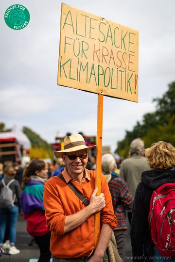Ein älterer Mann steht inmitten eines Demonstrationszuges und schaut grinsend in die Kamera. Er trägt einen orangen Pulli, Sonnenbrille und Strohhut. An einer Stange hält er ein Schild hoch, auf dem steht: Alte Säcke für krasse Klimapolitik!