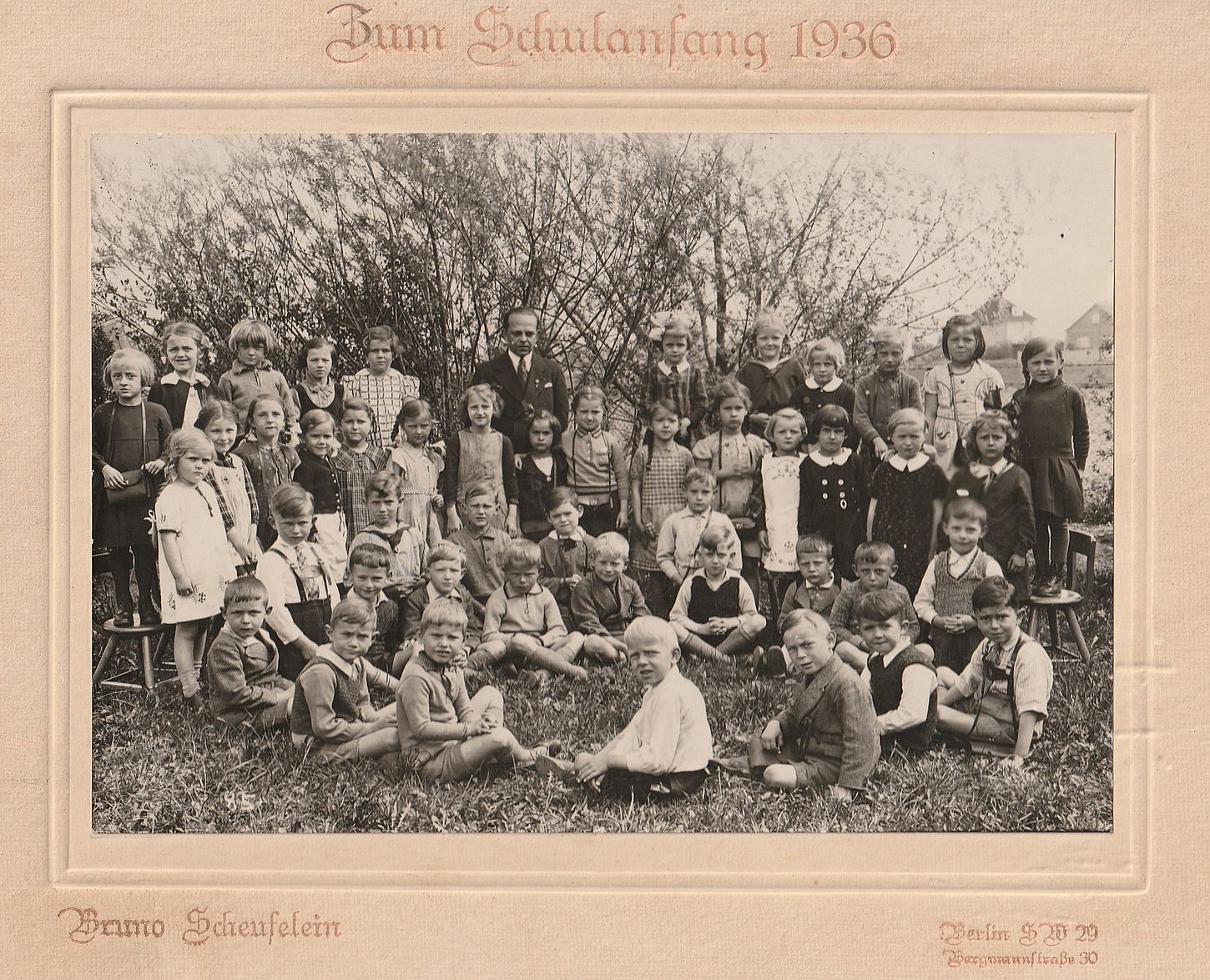 Eine Gruppe von 48 Kindern, im Gras, vor einem Gebüsch. Im Hintergrund sieht man vereinzelte Häuser. Vorne sitzen am Boden die Jungs, die Mädchen stehen hinten, in der hinteren Reihe auf Stühlen. In der Mitte hinten steht ein formal gekleideter, streng blickender Mann. Es sind etwas mehr Mädchen als Jungs. Kaum eines der Kinder lächelt.