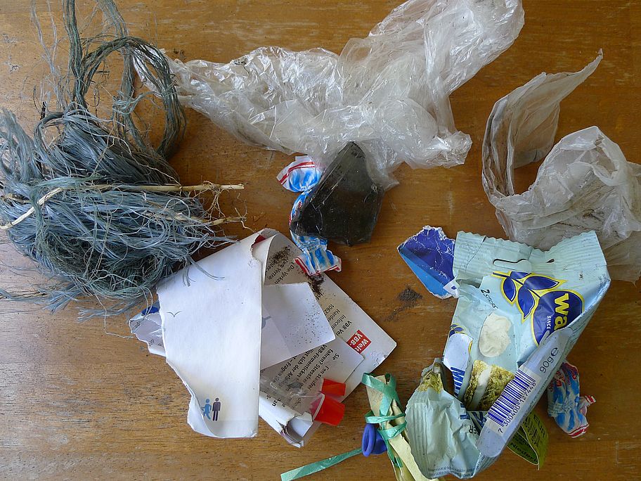 Auf einem Tisch liegt ein Haufen Abfall: viele Papierstücke, ein zerfasertes, dickes Bündel grauer Kunststoffschnur, eine große Glasscherbe, Plastiktüten, Bonbonpapiere, ein Luftballon mit einem Stück Schnur und eine Knäckebrotverpackung.