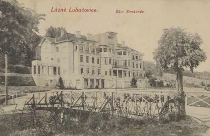 Ein recht großes, vierstöckiges Gebäude. Mit verschiedenen Giebeln und Balkonen, in freier Landschaft oder einem Park gelegen. Im Vordergrund eine kleine Brücke.
