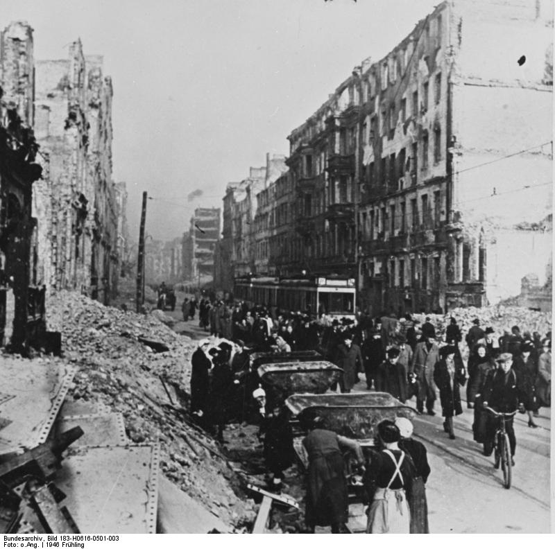 Eine schmale Straße, gesäumt von hohen mehrstöckigen Häusern, alle stark beschädigt. In der Straßenmitte steht eine Straßenbahn, drumherum, davor und dahinter laufen viele Menschen, alle winterlich dunkel gekleidet. Die ganze Straße entlang stapelt sich vor den Häusern der Schutt. Im Vordergrund sind einige Frauen damit beschäftigt, ihn in bereitstehende Loren zu schaufeln.