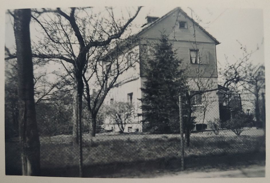 Ein schlichtes zweistöckiges Gebäude (EG + 1. Stock). Man erkennt auf jeder Etage an jeder Seite zwei Fenster und eins unter dem Giebel des Daches. Im Hintergrund ist ein weiteres Wohnhaus zu erkennen, ein ganzes Stück vor dem Haus ist ein Maschendrahtzaun, drum herum stehen Bäume, vermutlich Obstbäume.