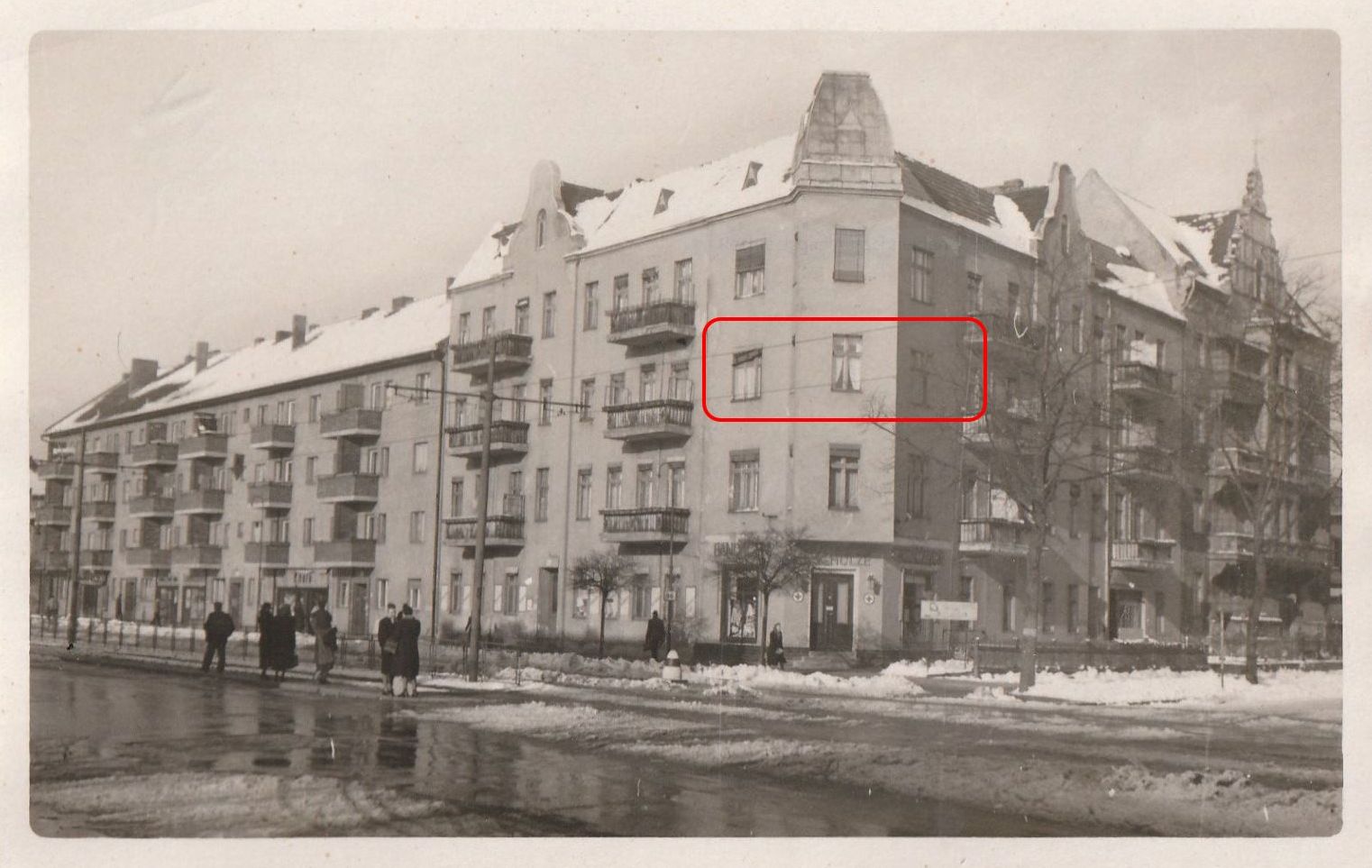 Ein großes, dreistöckiges Eckgebäude an einer großen Straße. Auf der Straße sieht man dunklen Schneematsch und an den Rändern weißen Schnee. Die Straße ist breit, aber es fahren keine Autos. Einige Menschen warten in der Mitte der Straße an einer Straßenbahnhaltestelle. Das Haus hat Balkone, im Erdgeschoss ist ein Laden, vor dem Haus stehen in der Hauptstraße kleine und in der Nebenstraße große Bäume, die aber alle unbelaubt sind.