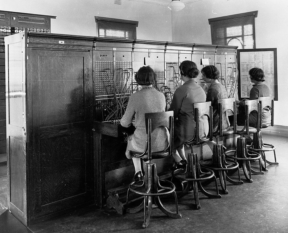 Vier Frauen sitzen auf hohen Stühlen vor einem großen, hölzernen Schrank. Darin sind vor ihnen zahlreiche Löcher, vor jeder der Frauen sind einige der Löcher durch eingesteckte Kabel verbunden. Die Frauen tragen Kopfhörer, ein Stuhl ist frei.