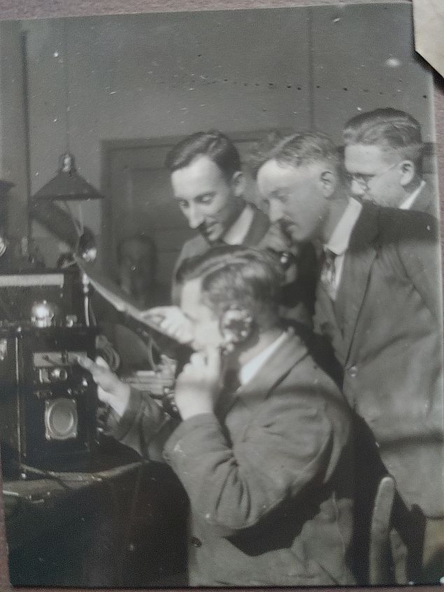 Vier Männer um einen großen alten Telefonapparat herum. Einer sitzt um hält den Hörer ans Ohr, die anderen stehen dicht dabei und schauen interessiert zu. Der Apparat ist groß und schwarz und wirkt sehr technisch, oben drauf leuchtet eine Lampe. Die Männer tragen alle Anzug und Krawatte.