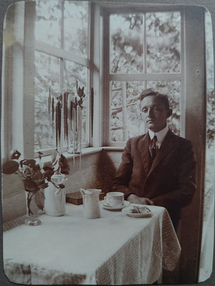 Ein Mann sitzt an einem kleinen Tisch in einem Glasanbau, neben einer offenen Tür. Auf dem Tisch stehen eine Kaffeekanne, eine Tasse und ein kleiner Teller mit Gebäck. Er ist ordentlich gekleidet, dunkler Anzug mit steifem Hemdkragen und Krawatte. Er blickt nicht in die Kamera und lächelt nicht, er sieht melancholisch, ja traurig aus und schaut ins Leere.