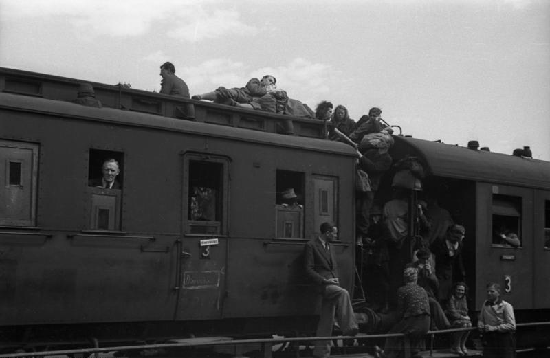Ein Blick auf die Verbindung zwischen zwei Eisenbahnwaggons. Der Zwischenraum auf den Puffern ist dicht besetzt und auch auf dem Dach sitzen schon einige Menschen.